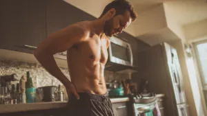 Muscular man in kitchen checking his abs, showcasing fitness and physique, emphasizing health, supplements, and wellness.