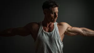 High-quality image of a muscular man in a white tank top showcasing defined arms and physique, emphasizing fitness and strength with a dark background.